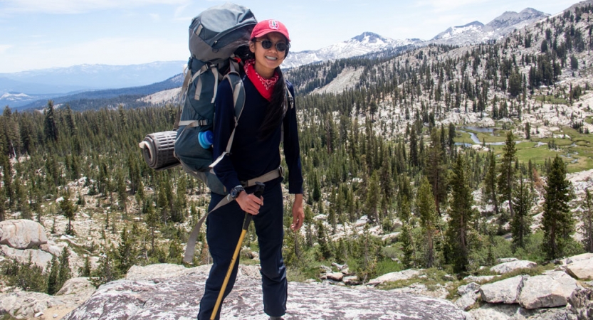 a person carrying a backpack smiles at the camera with a mountain landscape in the background 